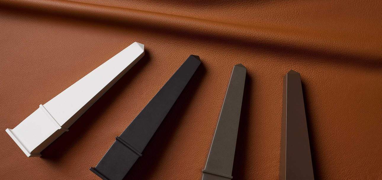 Close-up of Foglizzo leather, rich cognac-colored textured leather backdrop featuring four tall, pointed aluminum McKinnon and Harris finish obelisks resting on top in Classic White, Sorghum Black, Natural Bronze, and Jura Peat.