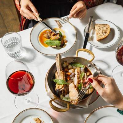 Overhead view of an elegant dining setting with a copper serving pan, a plated appetizer, sliced bread, a glass of red wine, and hands holding utensils.