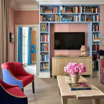 A stylishly decorated hotel suite centered around a built-in blue bookshelf framed around a flat-screen television, featuring vibrant armchairs and floral accents.