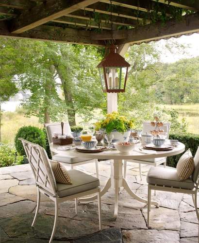 Outdoor dining area featuring a McKinnon and Harris white round dining table set for breakfast under a wooden pergola, overlooking a lush green landscape and pond. The table is surrounded by four Otey dining chairs with light grey cushions and striped throw pillows. A rustic lantern hangs above the centerpiece of yellow flowers, and the setting is on a natural stone patio. Design by Cathy Kincaid, Photo by Tria Giovan