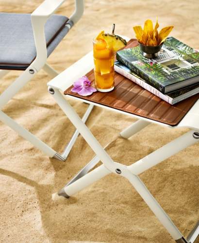 Close-up of McKinnon and Harris Scout Folding Table on sand with a tropical cocktail, and a book resting resting on Foglizzo leather pad.