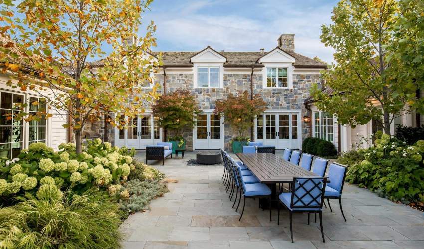 Elegant outdoor entertaining space in the fall, showcasing a long McKinnon and Harris Parsons dining table and Otey Dining Chairsfor large gatherings. The patio is framed by mature landscaping, a casual fire pit area, and the classic architecture of a stone-clad home. Design by Hillary Taylor Interiors, Photo by Lindsay Salazar