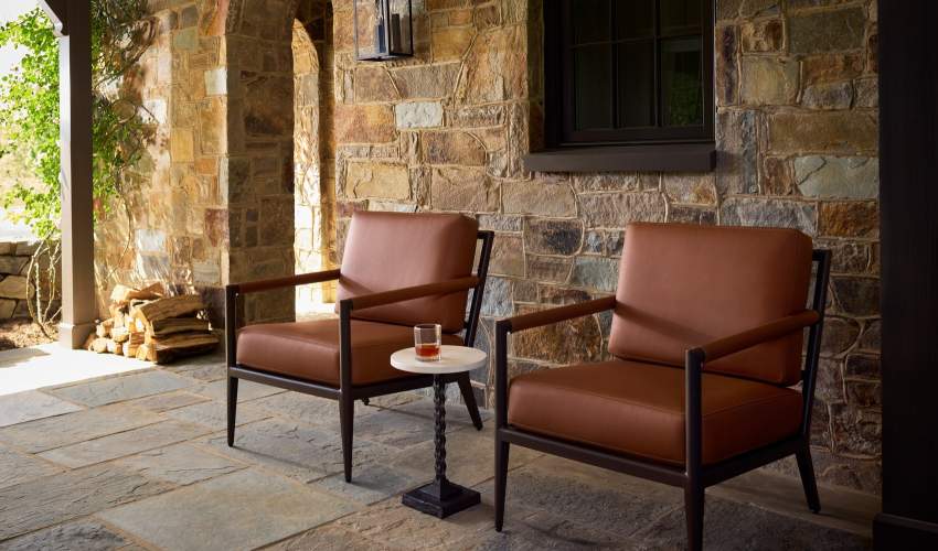 Two dark frame outdoor armchairs with Foglizzo brown leather cushions around a small white top side table on a stone patio with a stone wall background.