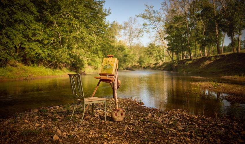 Buie Dining Side Chair with High Performance Rush Seat finished in Shenandoah Gray Green next to a creek with an art easel set up