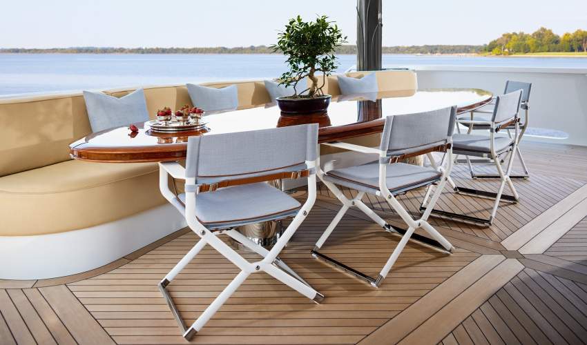 Outdoor dining setup on a yacht deck overlooking open water, featuring a oval dining table, classic white Scout folding director chairs with leather back-straps, and built-in tan bench seating.